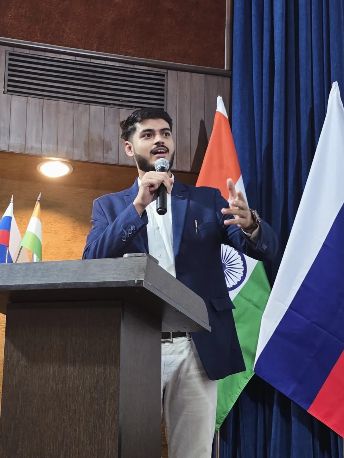 Akshat Mehrotra speaking at a podium with Indian and Russian flags.